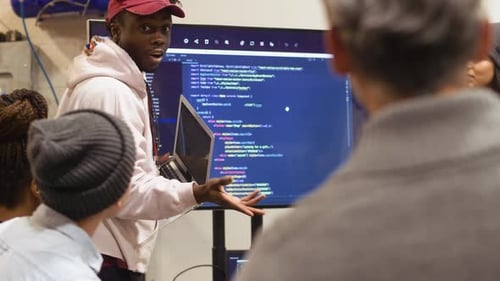 Young adults watch presentation with code on monitor.