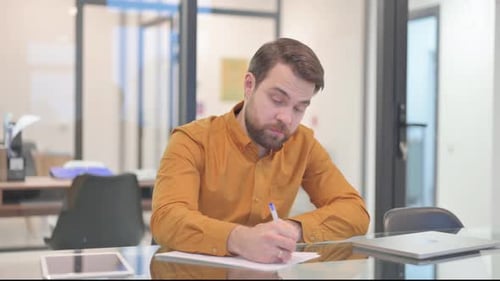Bearded Man Writing at Desk in Modern Office