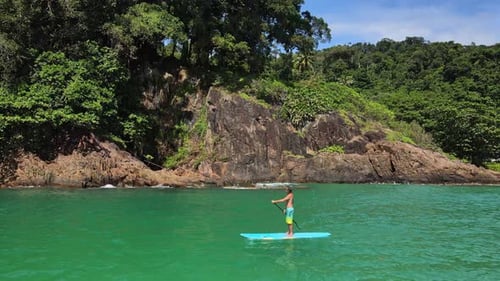 Trucking aerial drone shot of Caucasian man exercising on a sup paddle board in turquoise tropical c