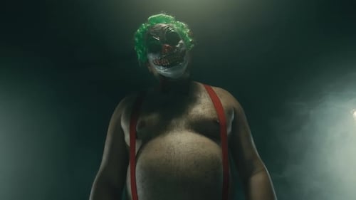 Scary Man in the Horror Clown Mask with Green Hair Standing in Suspenders in the Dark Studio with
