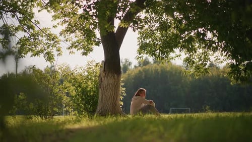 Woman Sitting Under Tree in Sunlit Park Surrounded By Greenery and Gentle Wind