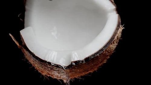 Coconut milk or juice is poured from the half with white flesh, close-up on a black background. Slow