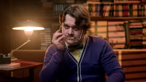 Un homme moustachu sérieux en vêtements rétro est assis dans une bibliothèque vintage et met des lunettes, portrait élégant
