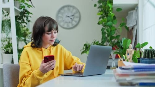 Young Female Sitting at Table at Home Using Smartphone and Laptop