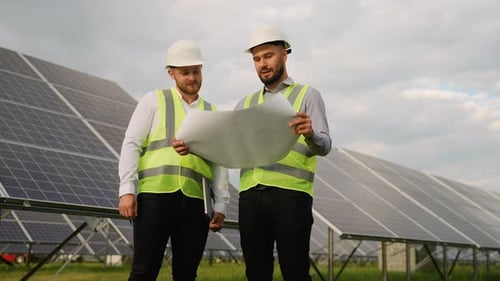 Two Male Engineers in Safety Vests and Hard Hats Discussing Blueprint at Solar Panel Farm