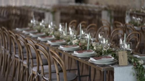 Long, elegantly set wedding tables with floral centerpieces and wooden chairs in a reception hall.