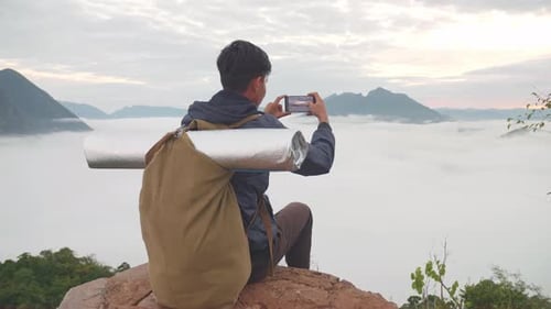 Homem caminhante sentado na rocha e usando smartphone tirando foto da vista superior da montanha nebulosa