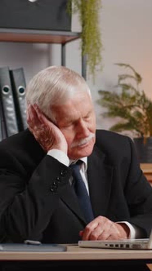 Bored Sleepy Senior Businessman Working on Laptop Computer Yawns Leaning on Hand at Office Desk