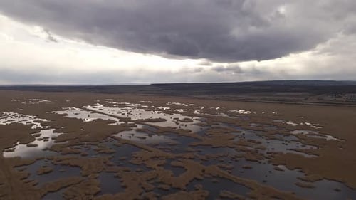 An aerial view of a vast wetland with patches of water reflecting the moody sky above. Dark clouds c