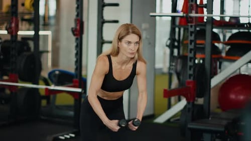 Concentrated Fit Middle Aged Lady Exercising with Dumbbells at Gym Leaning Forward and Lifting