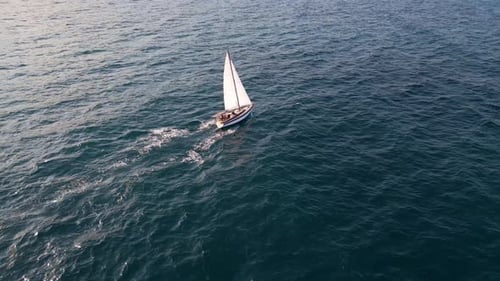 Aerial video of a white sailboat sailing alone on deep blue sea. Calm waves shimmer under sunlight.