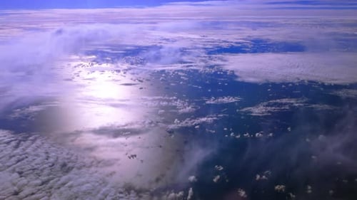 A high altitude aerial view over clouds and ocean on a sunny day, taken from the cabin of a plane.