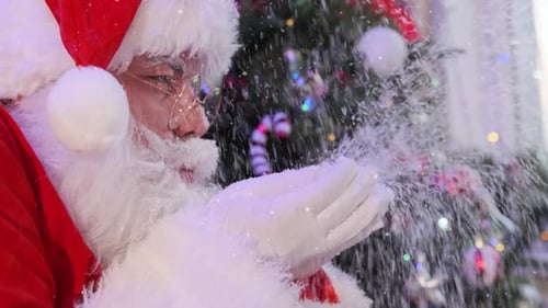 Close-up face of Santa Claus blowing flickering snowflakes from palms and looking into camera