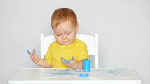 Concentrated Toddler Girl in Yellow Tshirt Touches Paint From Blue Jar and Draws with Fingers