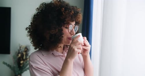 Attractive Peaceful Dreamy Woman with Curly Hair in Glasses Holds Cup and Enjoys Favorite Coffee
