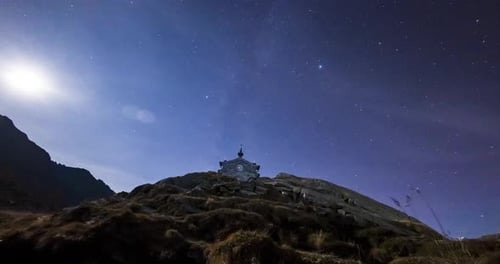 Timelapse of a serene mountain chapel under a starry night sky with the moon illuminating the landsc
