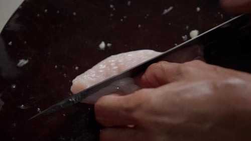 A person slicing raw chicken breast on a wooden cutting board, close-up