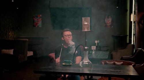 A man in glasses smokes a traditional hookah pipe A man exhales thick smoke in a hookah cafe or bar