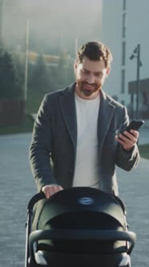 Father with Stroller Dad in Fashionable Coat Manages Stroller While Using Smartphone During Daily