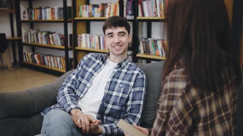 Young Man Engages in Conversation with Woman While Sitting on a Couch in a Cozy Library Filled with
