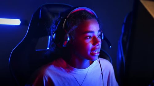 Black teen girl in headset playing video games in video game club with blue and red illumination, ta
