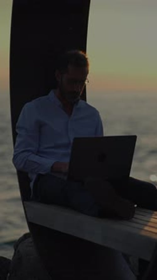 A Man is Working on His Laptop While Enjoying the Ocean View During a Beautiful Sunset