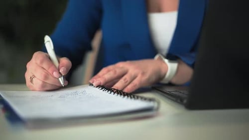 Young Woman Makes Notes in a Notebook with a Pen While Working at Computer in Office Closeup
