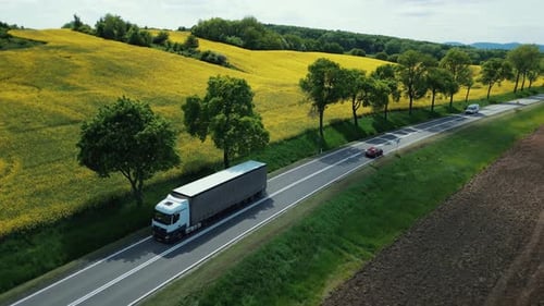 Aerial view truck and trailer driving on a scenic country highway along a flowering yellow field.