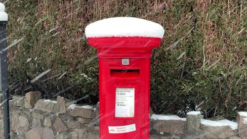 A British red post box for mail. It is snowing and the mailbox is covered in snow.