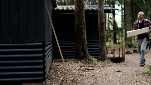 Man carrying wooden plank through forest path beside rustic cabins with copy space