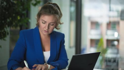 Cute Female Employee Working on a Laptop and Making Notes in a Notepad While Sitting in Home Office