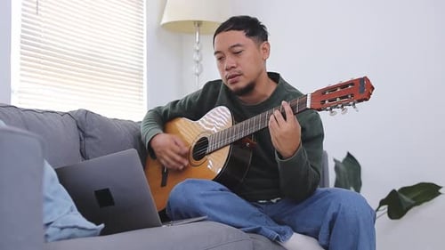 Man plays acoustic guitar while sitting on sofa