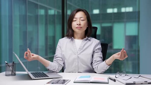 Asian businesswoman meditates with her eyes closed sitting at workplace in modern business office.