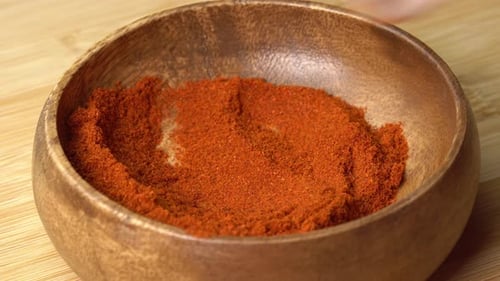 Spoon picks up Paprika powder spice from wooden bowl and rotating on wooden board. Dry seasoning