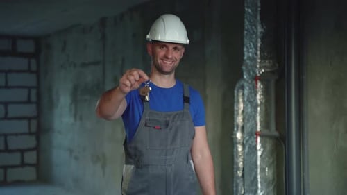 Glücklicher Handwerker in Overall und weißem Helm hält die Schlüssel zu einem neuen Haus in seinen Händen