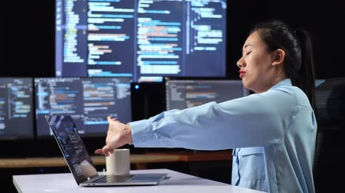 Side View Of Female Programmer Stretching While Writing Code By A Laptop Using Multiple Monitors