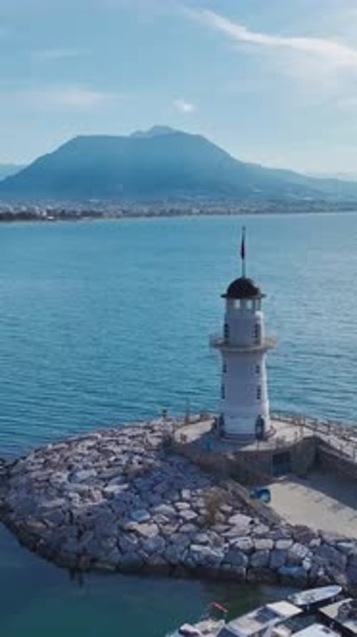Vertical Video of a White Lighthouse Surrounded By a Blue Sea Captured From a Drone With a Long Dock