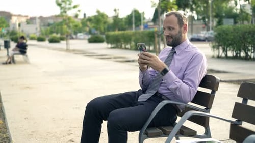 Businessman Texting On Smartphone Sitting On Bench In City