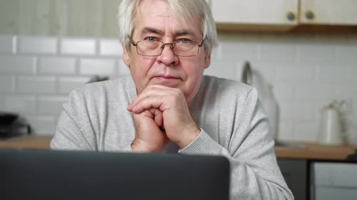 Portrait of Kind Old Man Sitting at Table with Laptop and Looking at Camera