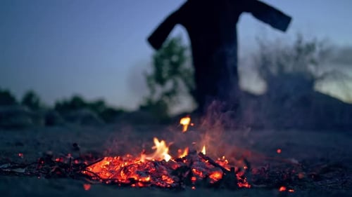 Embers of fire and scary witch outdoors. Death in black costume goes away from fire.