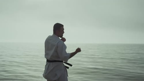 Starker Mann übt Taekwondo in der Nähe des Ozeans und macht aktive Handbewegungen. Sportlich muskulös