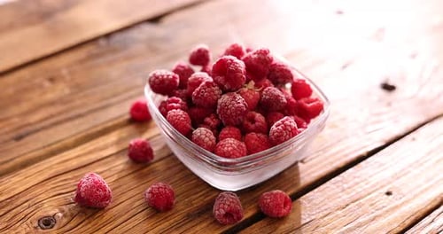 Ripe frozen raspberries and bowl on wooden table, closeup. Camera moving forward