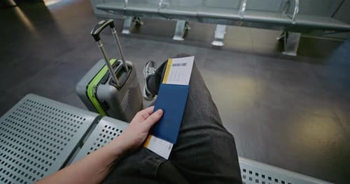 FirstPerson View of Traveler with Suitcase and Boarding Pass Sitting in Departure Lounge of Airline