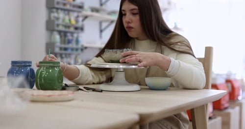 Creative young woman sitting at a table in a pottery studio, carefully painting a handmade ceramic b