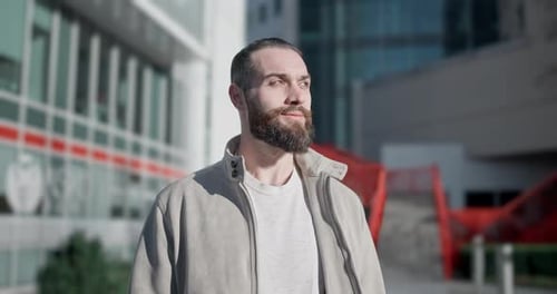 A Young Man with Stylish Trendy Hairstyle and Beard in Casual Clothes Stands on the Street in the