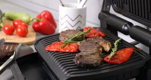 Elektrogrill zum Öffnen mit Rindersteaks, Rosmarin und Paprika am Tisch, Nahaufnahme. Kamerazoomen