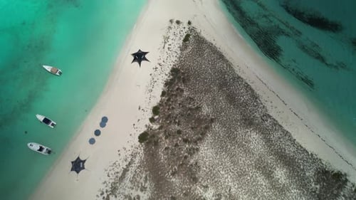 Boats and umbrellas on a sandy beach with turquoise waters and a grassy dune, aerial view