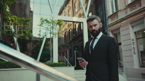 Serious Successful Business Man Uses Smartphone Go Away on the Big City Business District Street