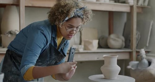 Young African American Woman in Apron Taking Photos of Ceramic Vase in Pottery Workshop Using