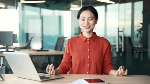 Young asian female employee meditates with her eyes closed sitting at workplace in business office.